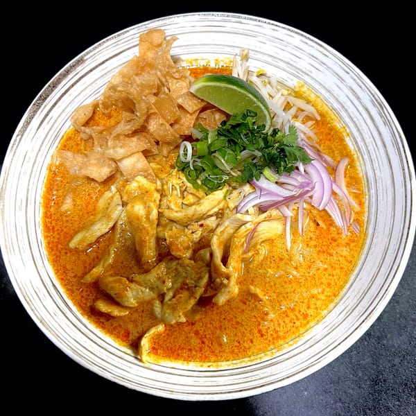 A bowl of Khao Soi with chicken, crispy noodles, red onions, cilantro, bean sprouts, and lime.