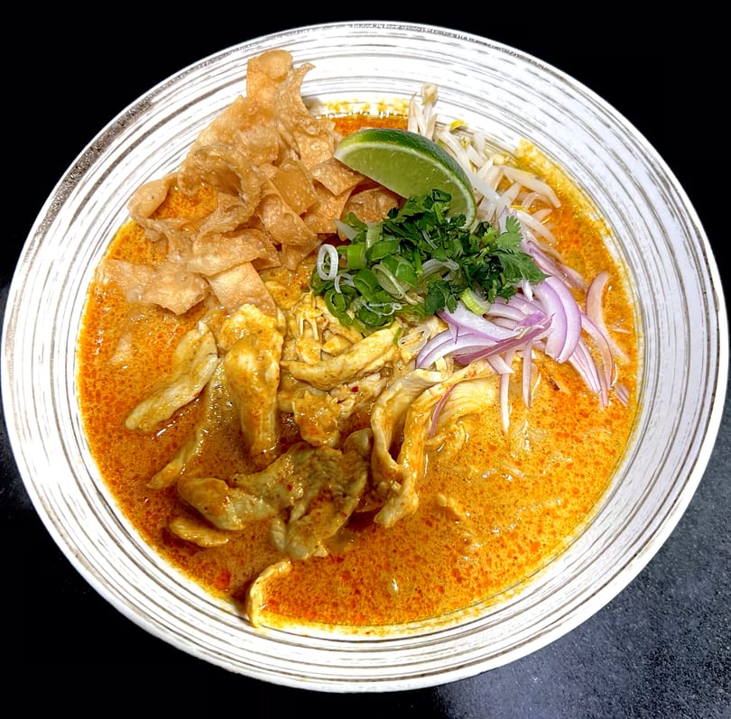 A bowl of Khao Soi with chicken, crispy noodles, red onions, cilantro, bean sprouts, and lime.