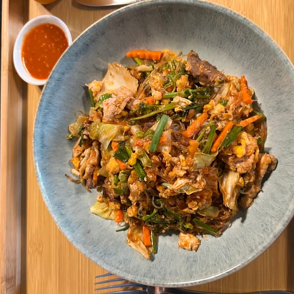 A blue bowl of sukiyaki stir-fry with meat, vegetables, and glass noodles, with a side of chili sauce.
