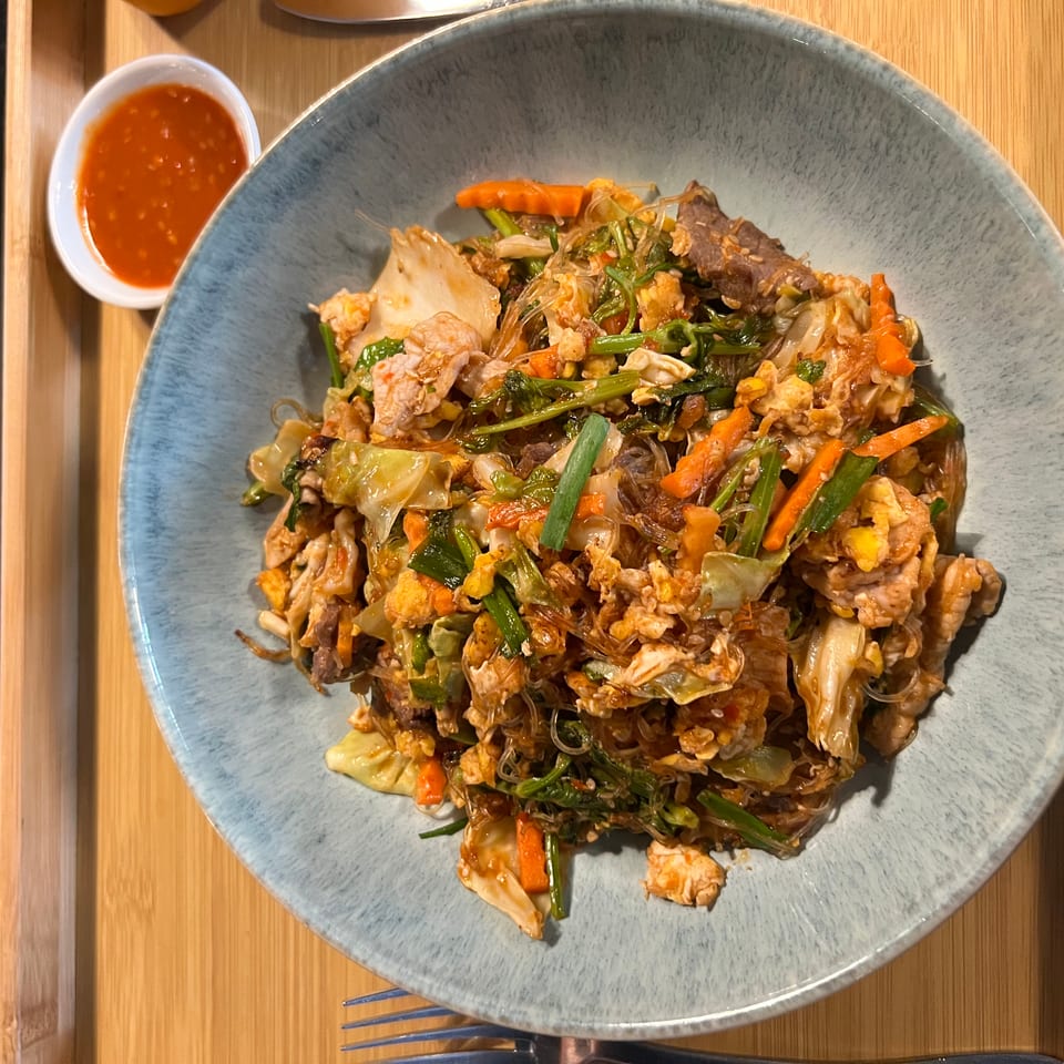 A blue bowl of sukiyaki stir-fry with meat, vegetables, and glass noodles, with a side of chili sauce.