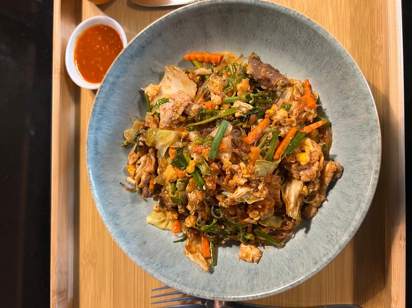 A blue bowl of sukiyaki stir-fry with meat, vegetables, and glass noodles, with a side of chili sauce.